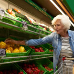 Portrait of modern senior woman with shopping cart choosing fruits and vegetables in supermarket while enjoying grocery shopping